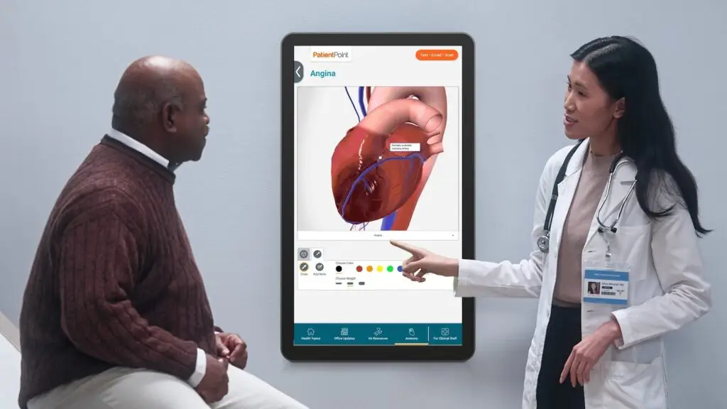Clinician in white coat points to a digital screen showing a 3D heart model labeled Angina while speaking with a patient.